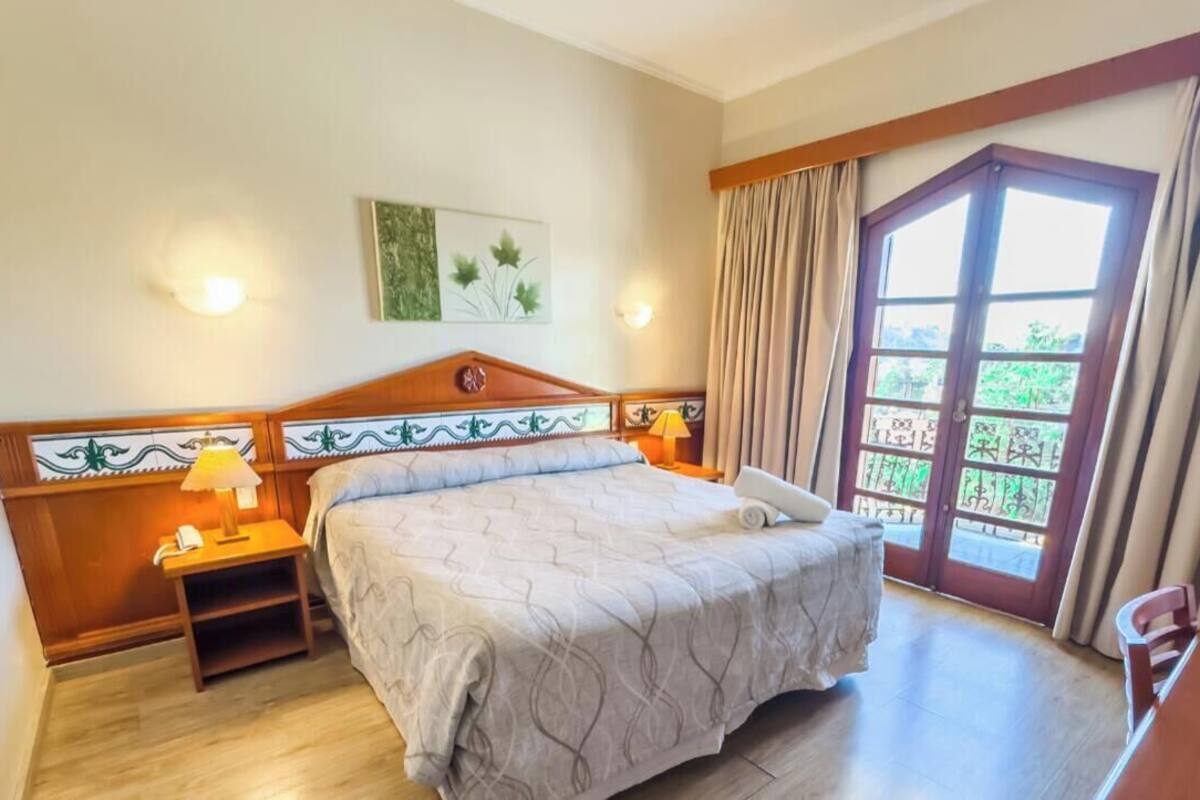 Habitación de hotel con cama, mesitas de noche, lámpara, teléfono y ventana con cortinas.