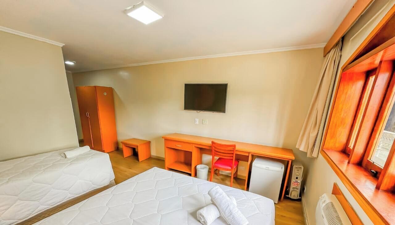 Quarto de hotel com duas camas, uma escrivaninha, uma cadeira, uma televisão e um guarda-roupa.