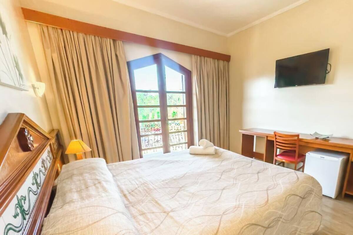 Una habitación de hotel con cama, escritorio, silla, televisor y ventana con cortinas.