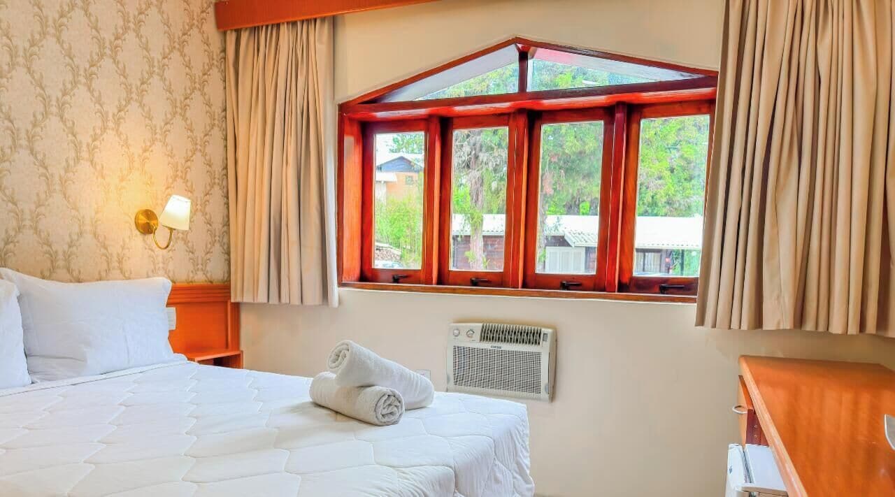 Una habitación de hotel con cama, ventana con cortinas, lámpara de pared y radiador.