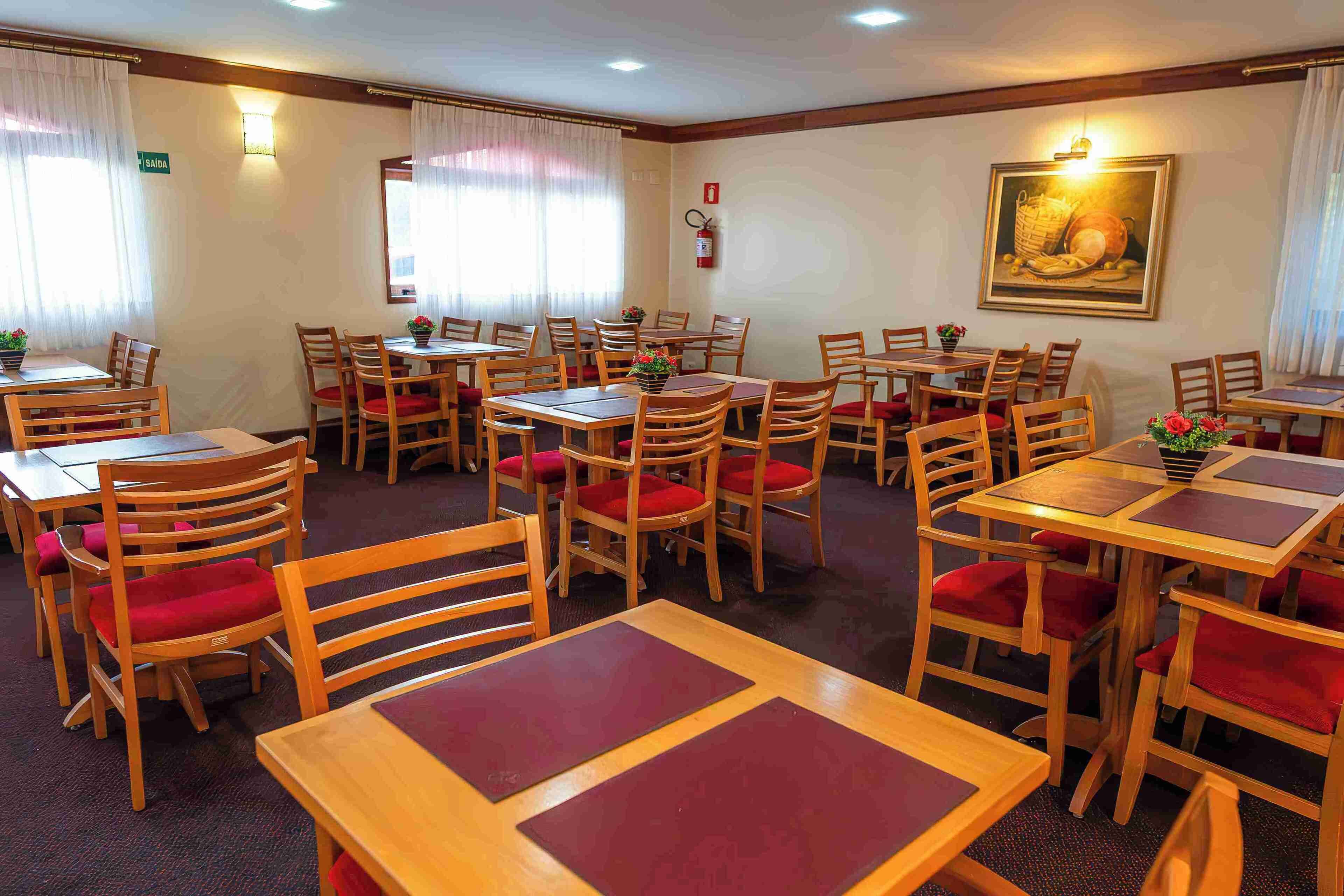 A hotel dining area with multiple wooden tables and chairs, some with red cushions. There are potted plants on the tables, and a fire extinguisher mounted on the wall.