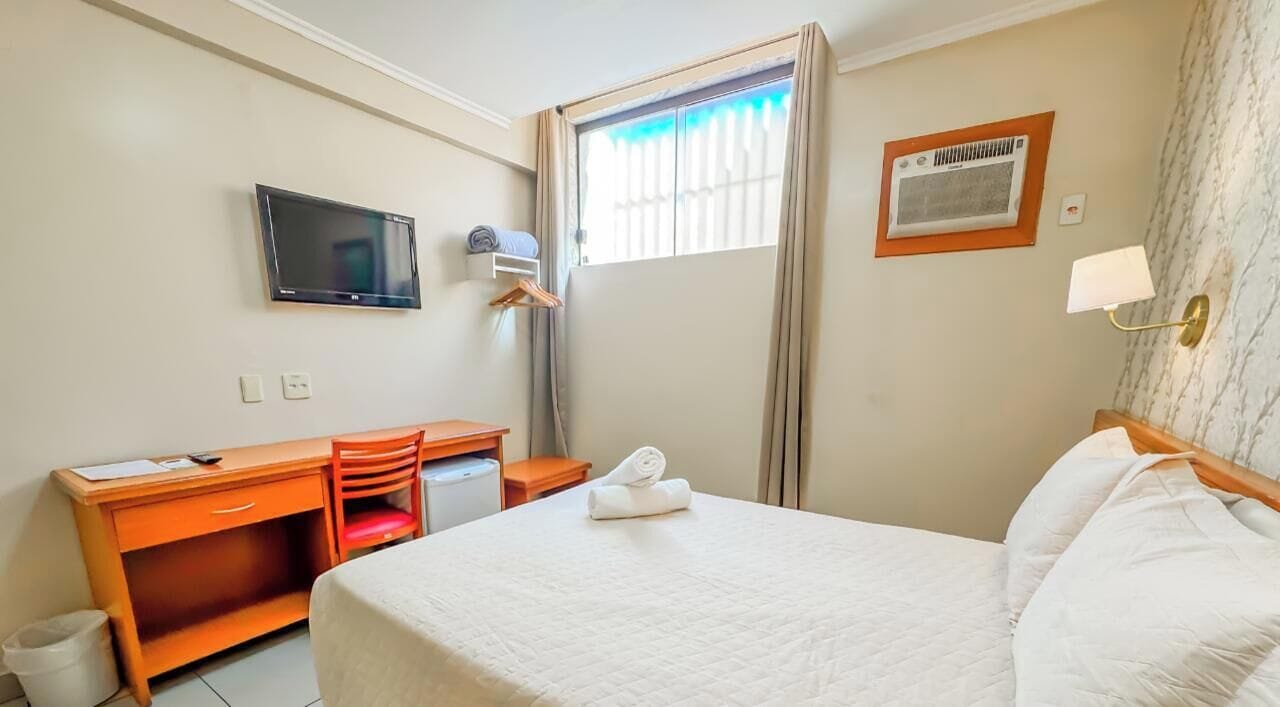 A hotel room with a bed, a desk, a chair, a television, a window with curtains, and a wall-mounted heater.