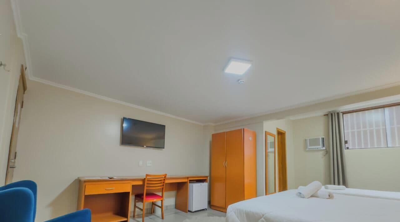 A hotel room with a bed, a desk, a chair, a wardrobe, a television, and a small refrigerator.