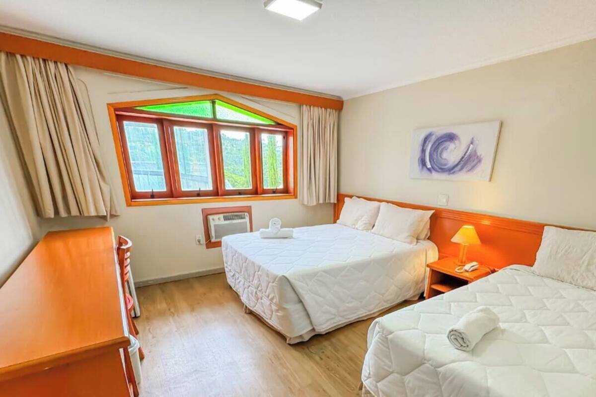 Habitación de hotel con dos camas, un escritorio, una ventana con cortinas, un radiador y un cuadro en la pared.