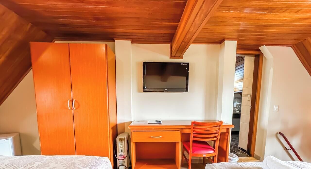A room with a wooden ceiling, a bed, a desk with a chair, a television, and a wardrobe.