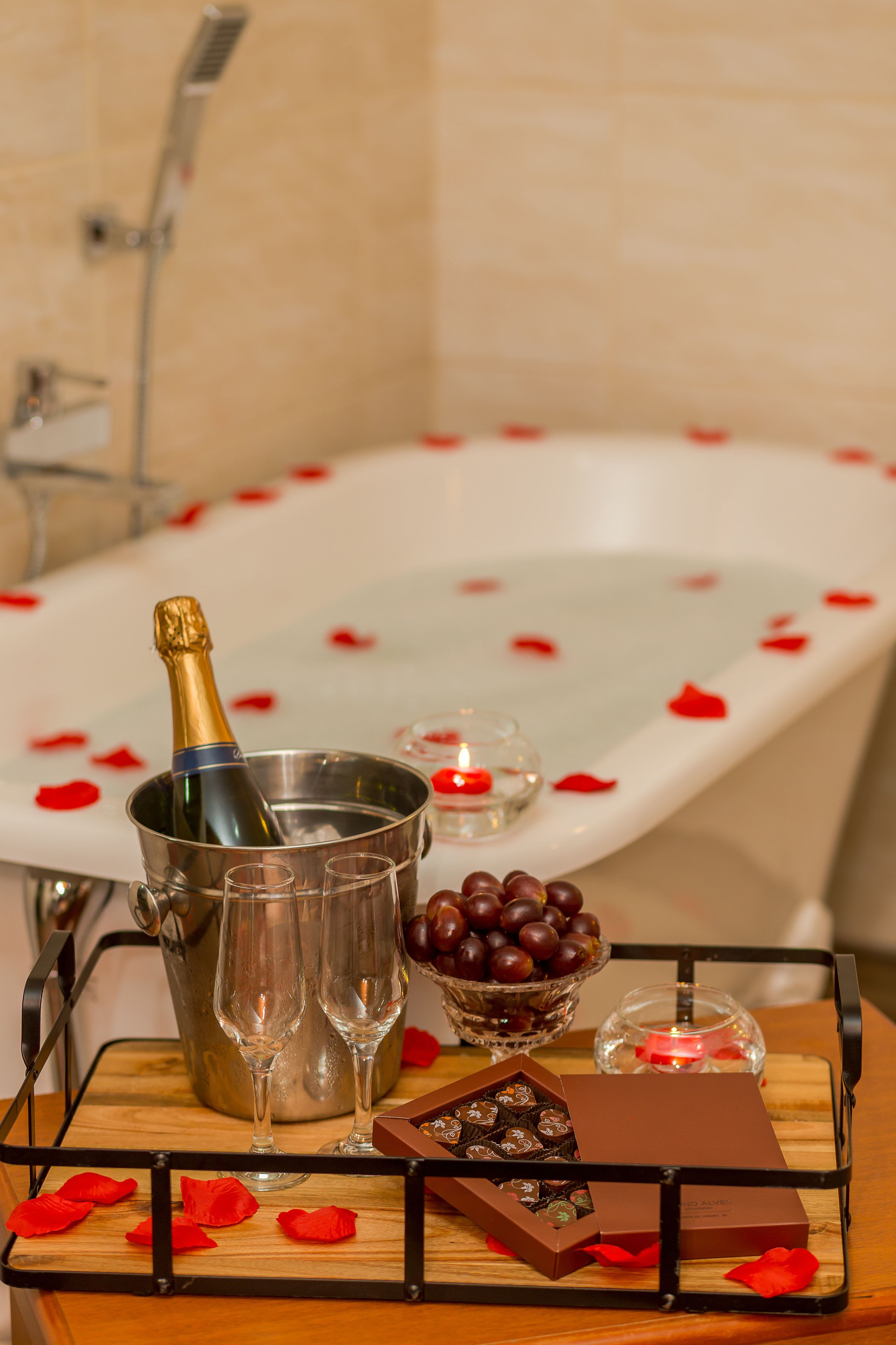A bathtub with rose petals, a champagne bottle, glasses, and chocolates on a tray.