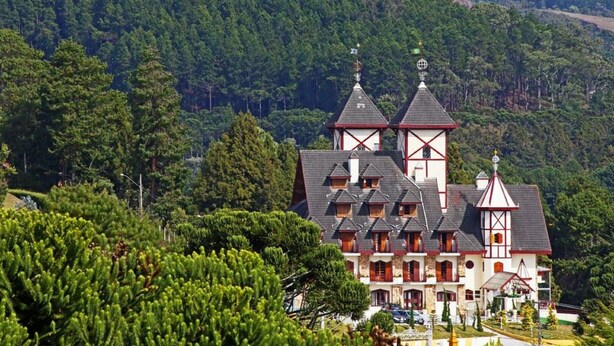 Un edificio de estilo chalé tradicional con múltiples techos a dos aguas y una torre destacada, rodeado de un bosque denso y colinas.