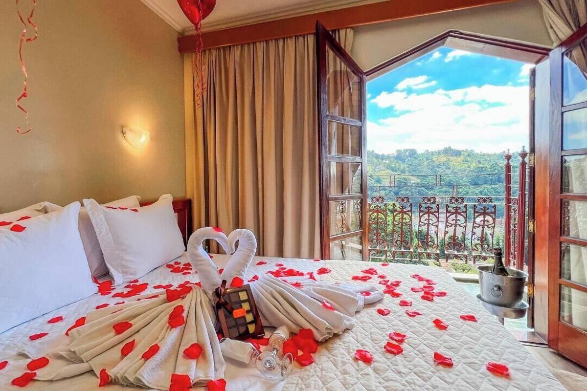 Habitación de hotel con una cama decorada con pétalos de rosa y una disposición de toallas en forma de corazón. Se observa un minibar en el lado derecho de la cama.