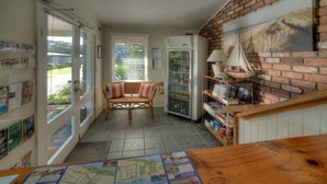 A cozy room with a wooden table, wicker chairs, a brick wall with a sailboat painting, a refrigerator, and a door leading outside.