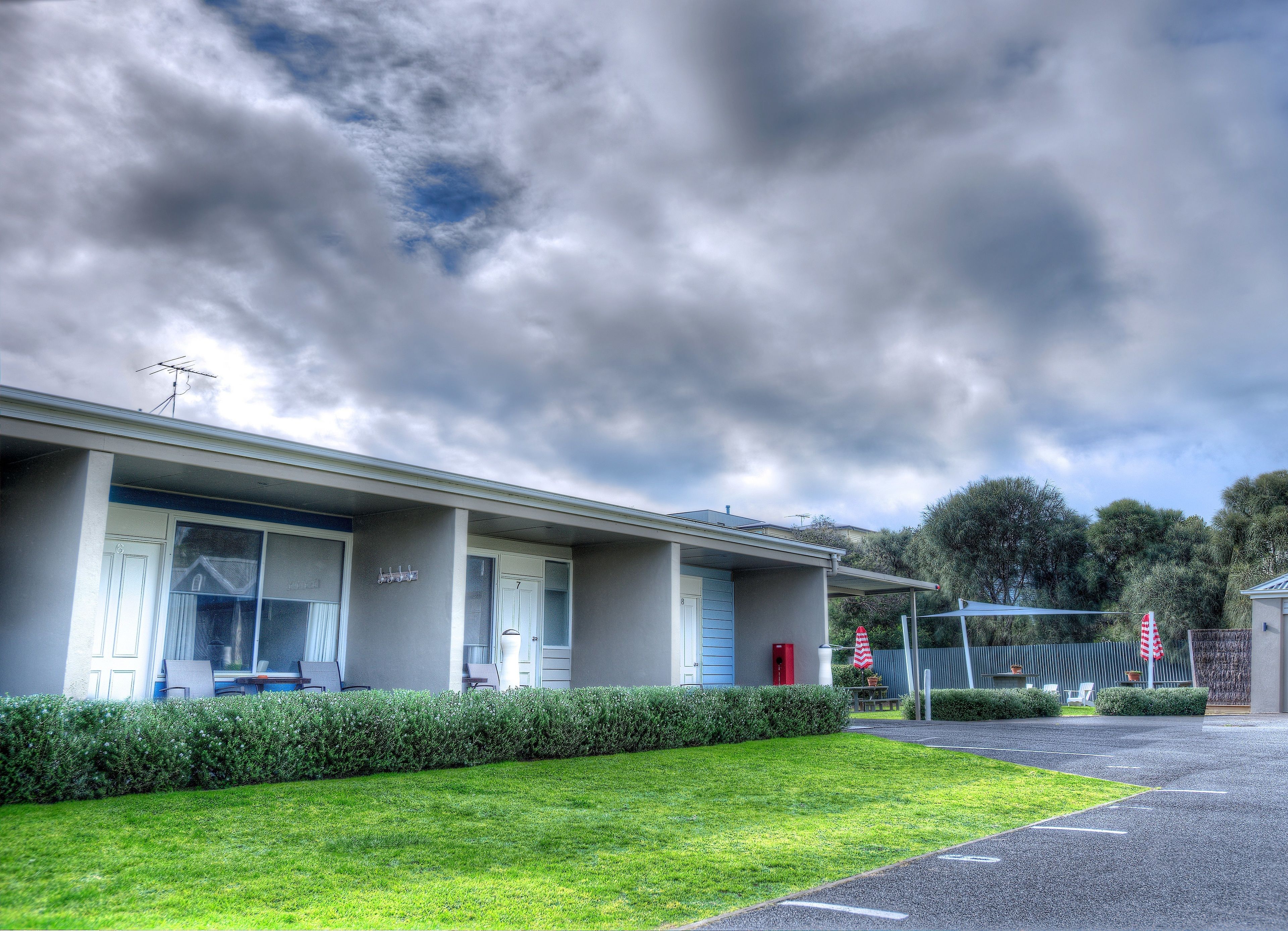 A motel with a covered parking area, a grassy lawn, and a row of rooms with a blue door.