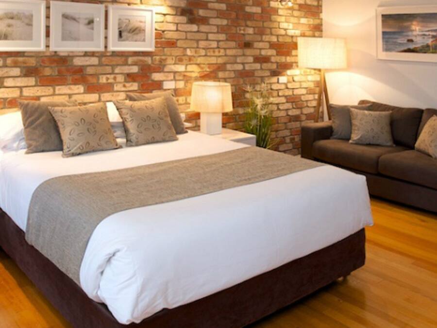 A bedroom with a brick wall, a bed with a white duvet, a grey sofa, and two bedside tables with lamps.