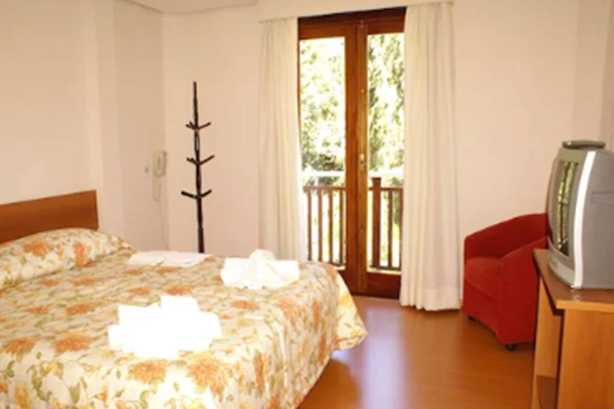 Un dormitorio con cama, una silla de madera, un televisor sobre un mueble y una puerta al balcón.