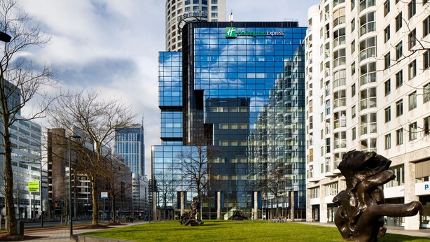 A modern hotel with a glass facade, surrounded by a grassy area and other buildings. - Holiday Inn Express Rotterdam - Central Station by IHG (Rotterdam)