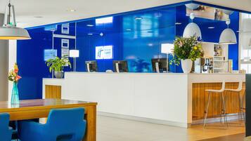 A hotel reception area with a blue accent wall, a reception desk, a seating area with blue chairs, and a potted plant.