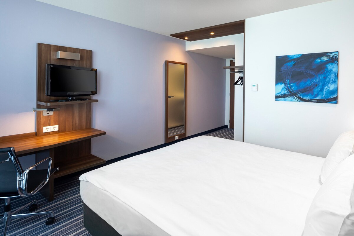 A hotel room with a bed, a desk with a TV, a chair, a painting, and a door.