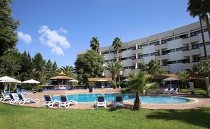 Une piscine entourée de palmiers et d’un immeuble à plusieurs étages, avec des chaises longues et des parasols.