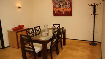 A dining area with a glass-top table, wooden chairs, a small cabinet with red bowls, a glass vase, and a painting on the wall.