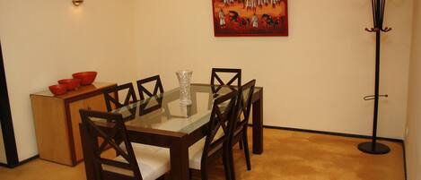 Un comedor con una mesa de vidrio, sillas de madera, un mueble pequeño con cuencos rojos, un florero de vidrio y un cuadro en la pared.