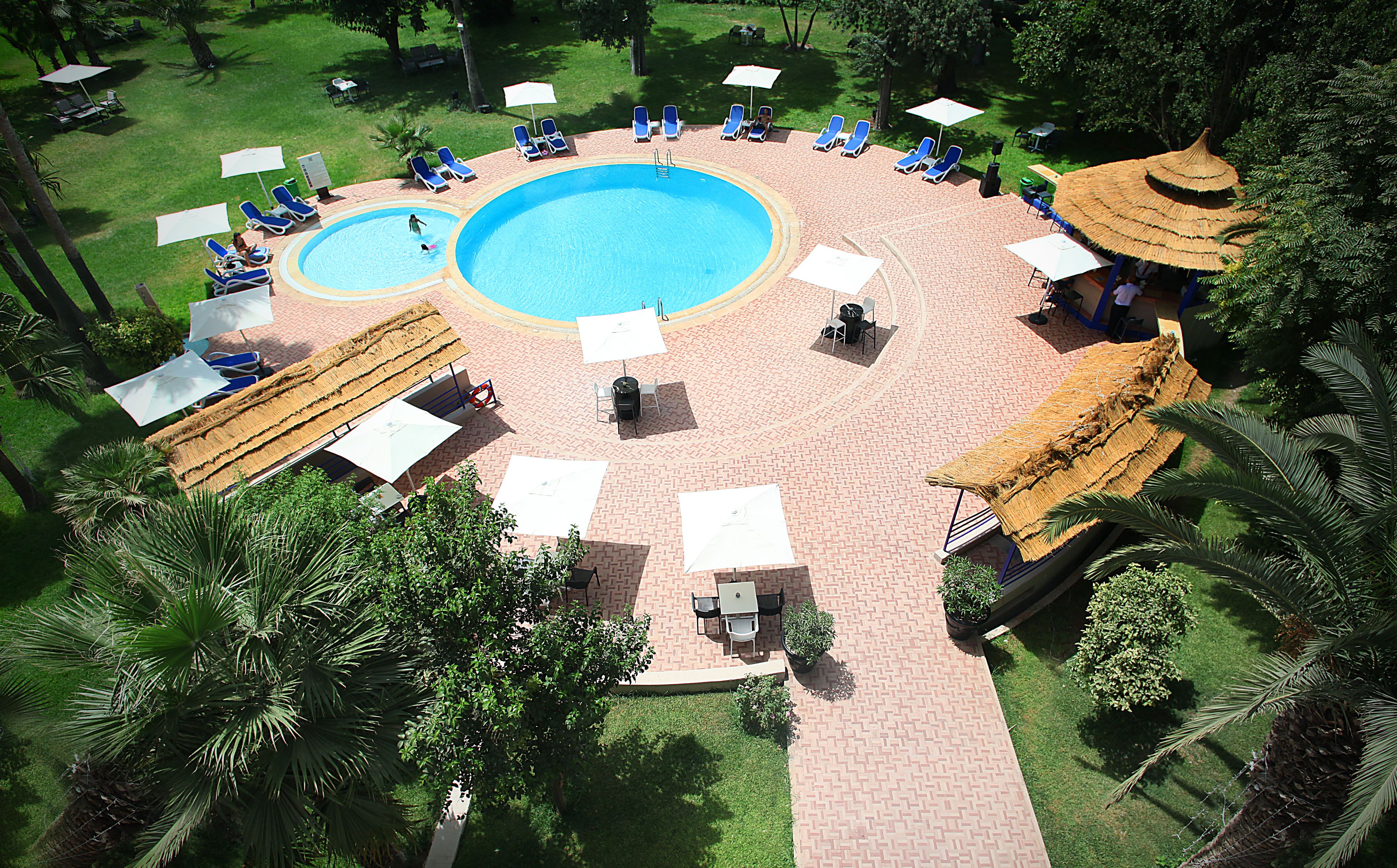 Ein kreisförmiger Poolbereich mit Liegestühlen und Sonnenschirmen, umgeben von einer Pflasterterrasse und Palmen.