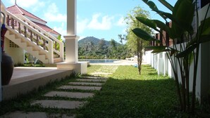 Un sendero conduce a una piscina con montañas al fondo, una escalera con techo rojo y un edificio blanco con columnas.