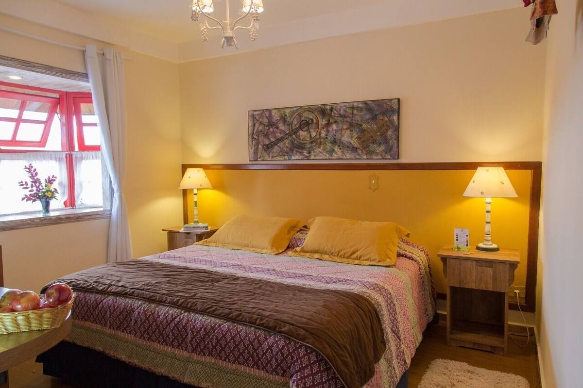 A bedroom with a bed, two bedside tables, a chandelier, a window with curtains, and a painting on the wall.