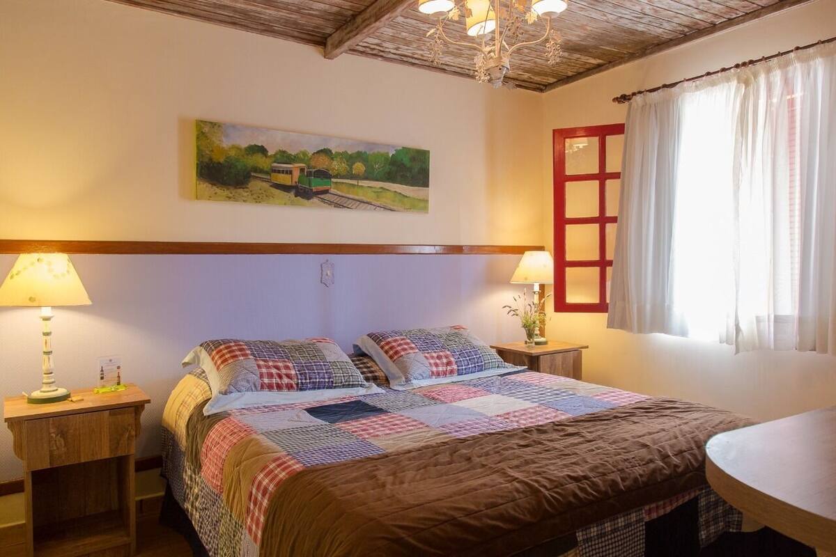 A bedroom with a large bed, two bedside tables, a chandelier, a painting on the wall, and a window with curtains.