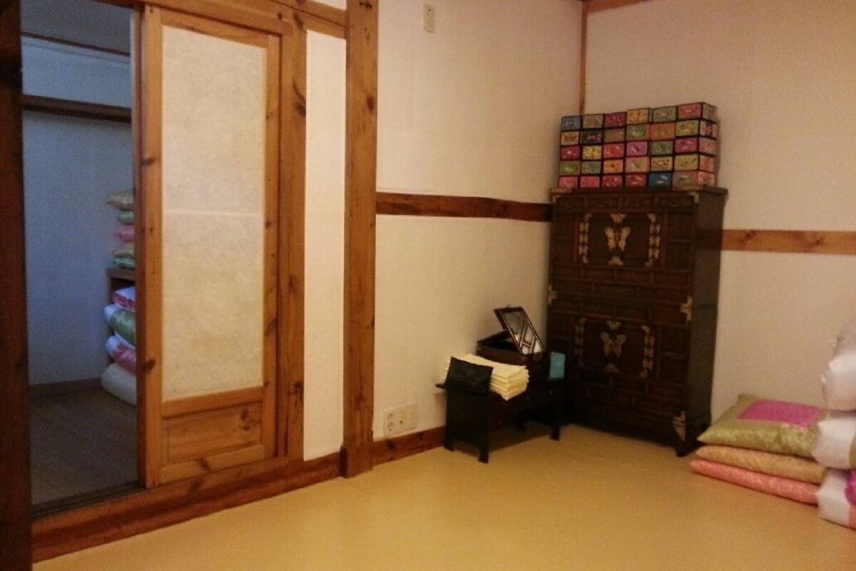 A room with a sliding door, a small table with a lamp, a cabinet with a box of games, and a stack of folded blankets.
