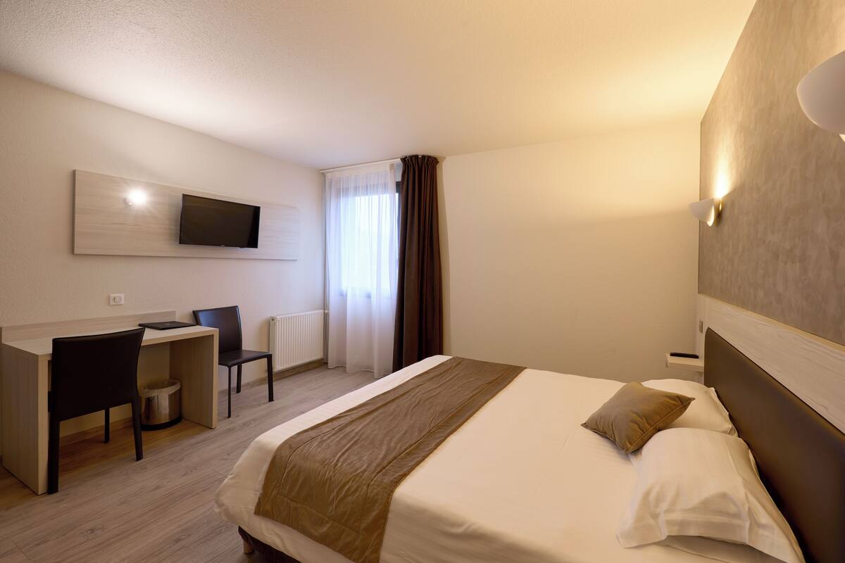 A hotel room with a bed, a desk, a chair, a television mounted on the wall, and a window with curtains.