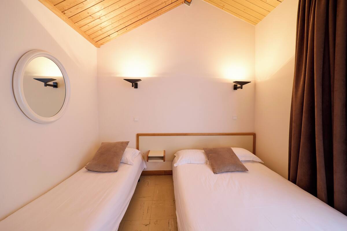 A small, well-lit room with two single beds, a round mirror, and two wall-mounted lights.