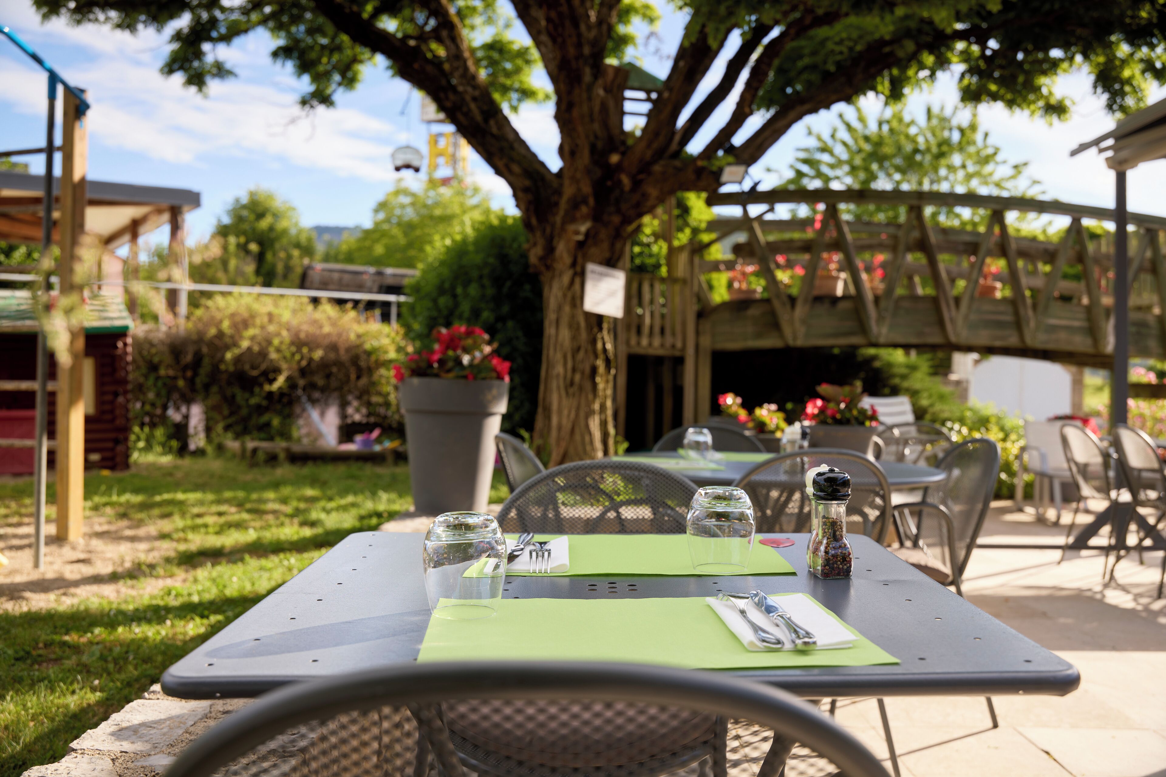 An outdoor dining area with tables set for two, surrounded by greenery and a tree.