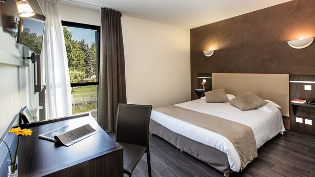 A modern hotel room with a large bed, a desk with a laptop, a chair, and a view of greenery outside. - Hotel Le Calice du Gevaudan (Banassac)
