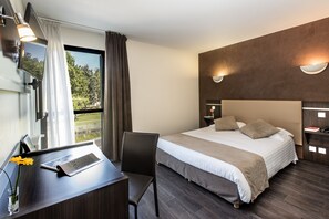 A modern hotel room with a large bed, a desk with a laptop, a chair, and a view of greenery outside. - Hotel Le Calice du Gevaudan (Banassac)