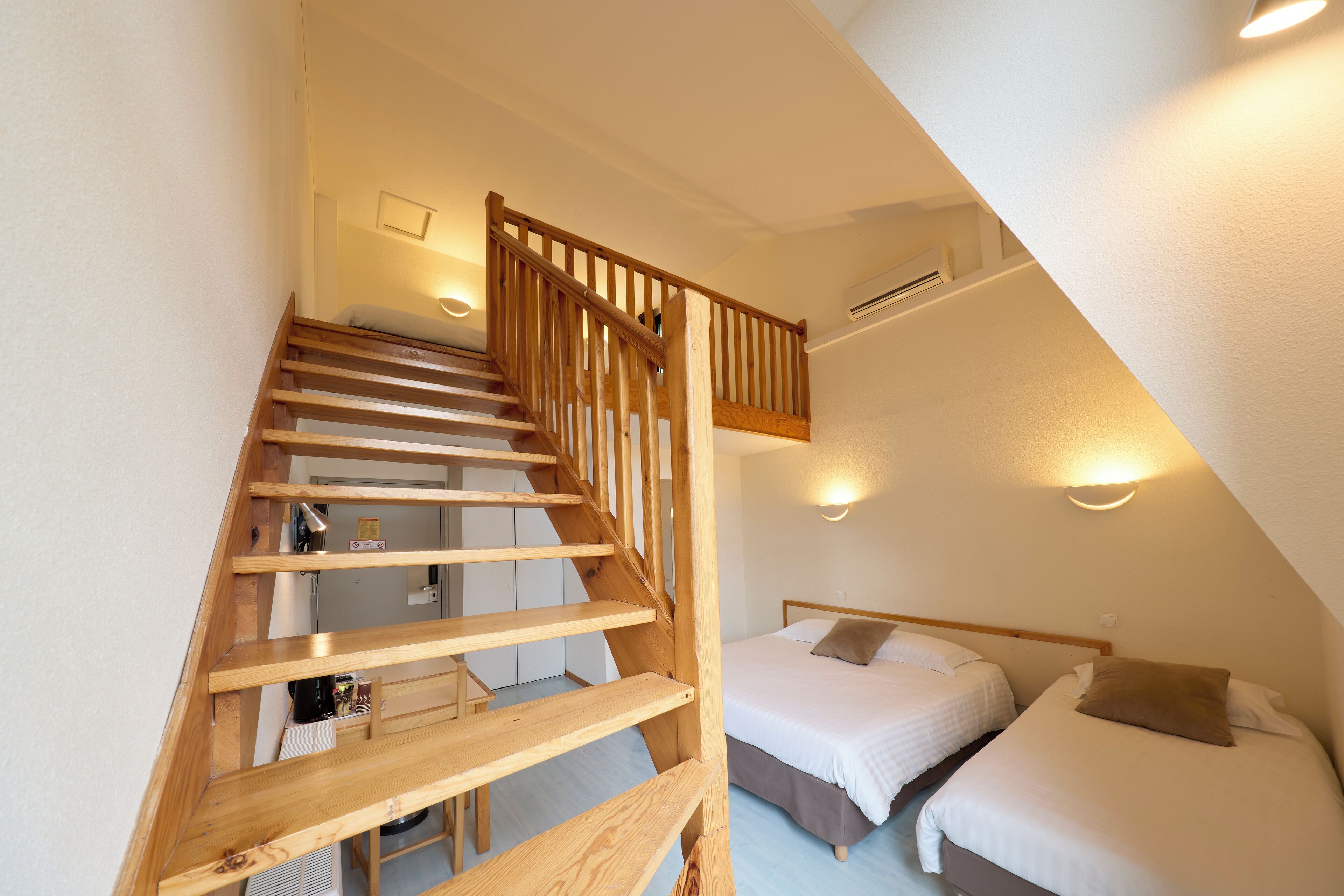 Un escalier menant à une chambre dotée de deux lits, d’une rambarde en bois et d’un luminaire au plafond.