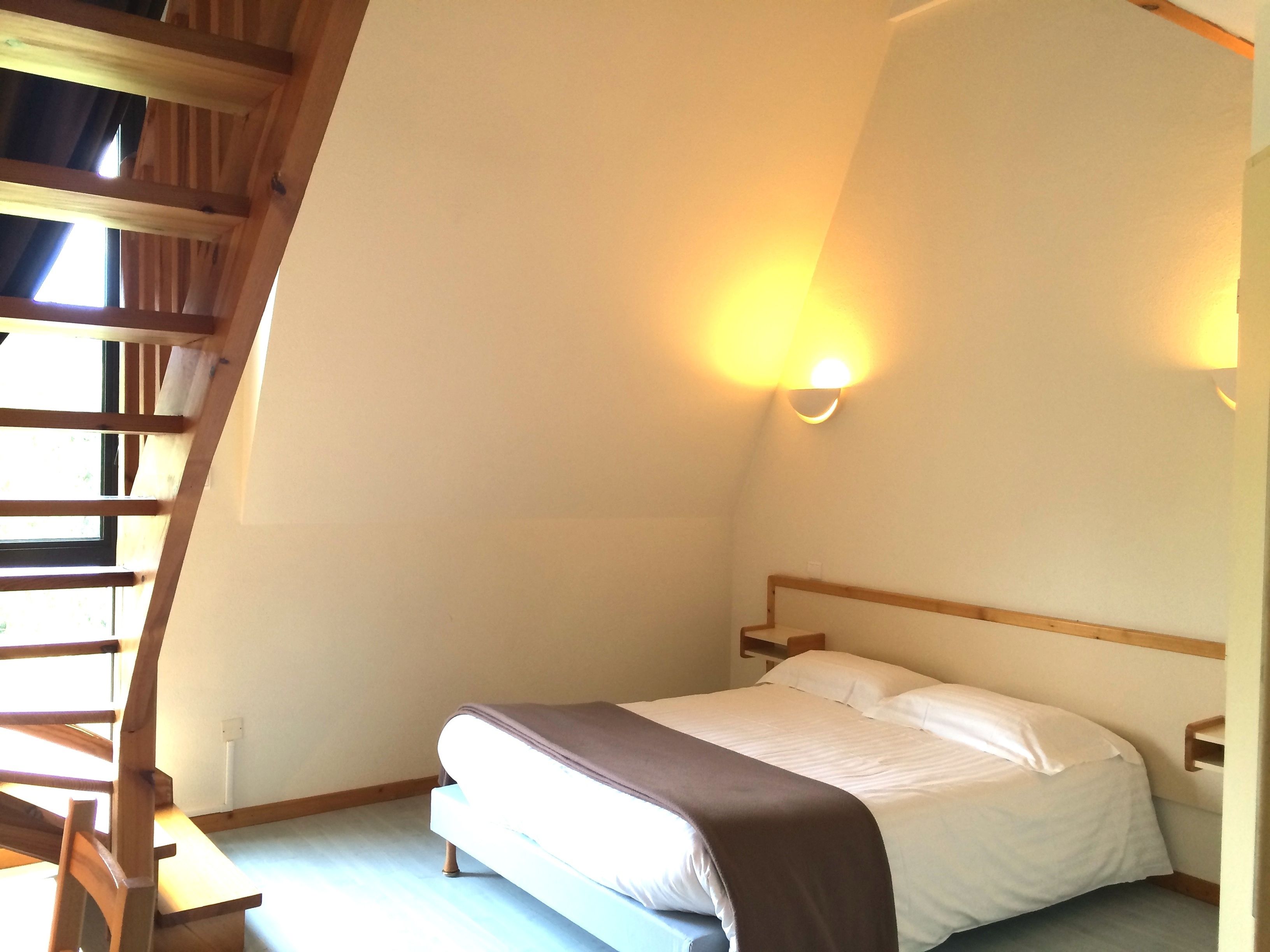 Un dormitorio con cama, cabecera de madera, mesita de noche, luz adosada a la pared y ventana con cortinas.