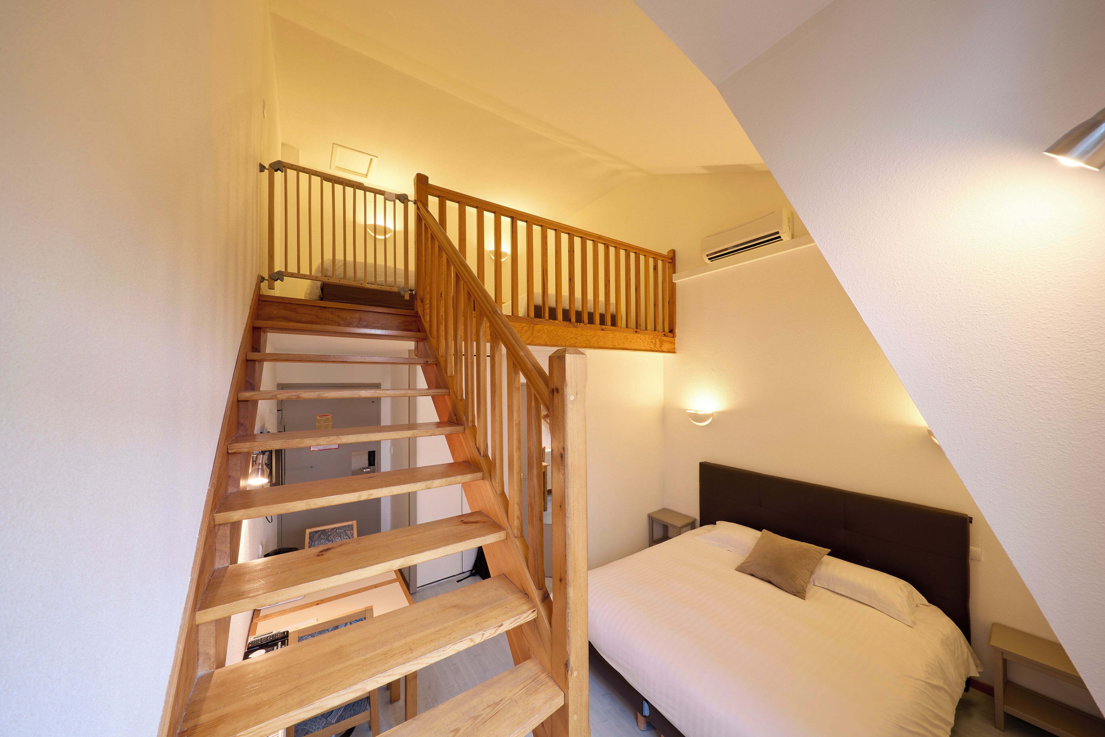 A bedroom with a wooden staircase, a bed, a nightstand, and a wall-mounted light.