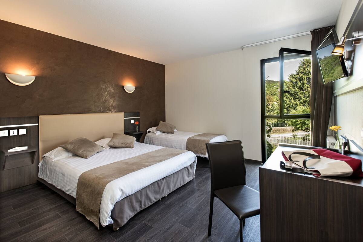 A modern hotel room with two beds, a desk, and a view of greenery.