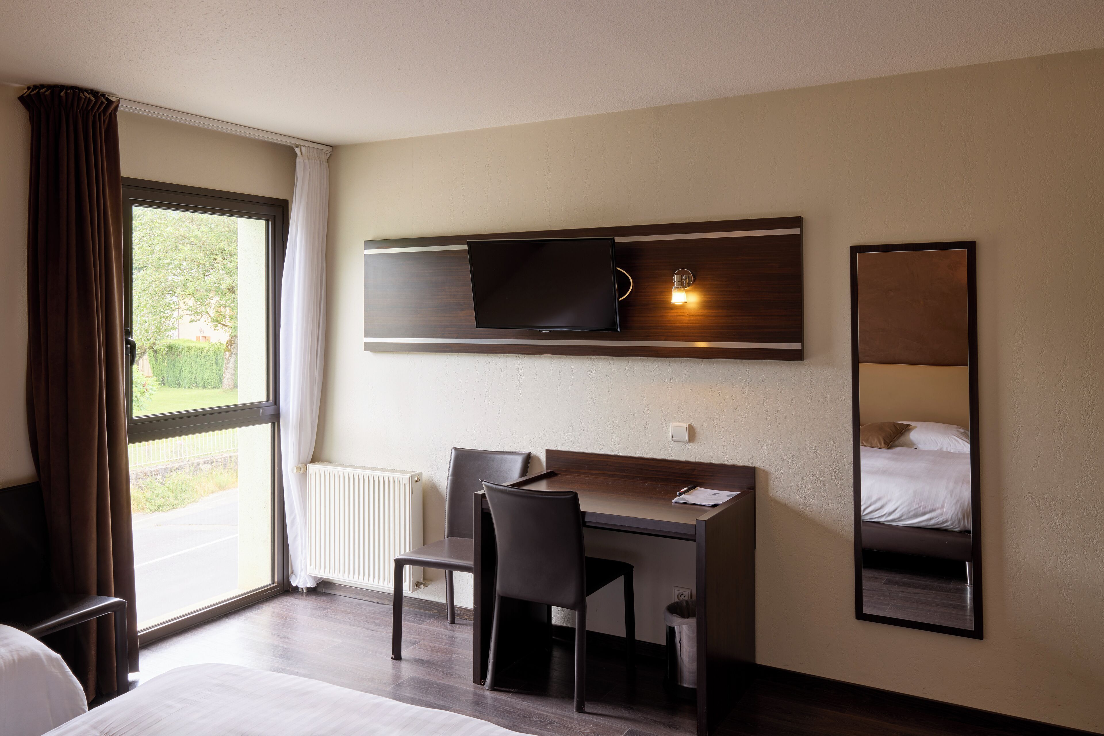 A hotel room with a bed, a desk with two chairs, a television mounted on the wall, and a large mirror reflecting the bed.