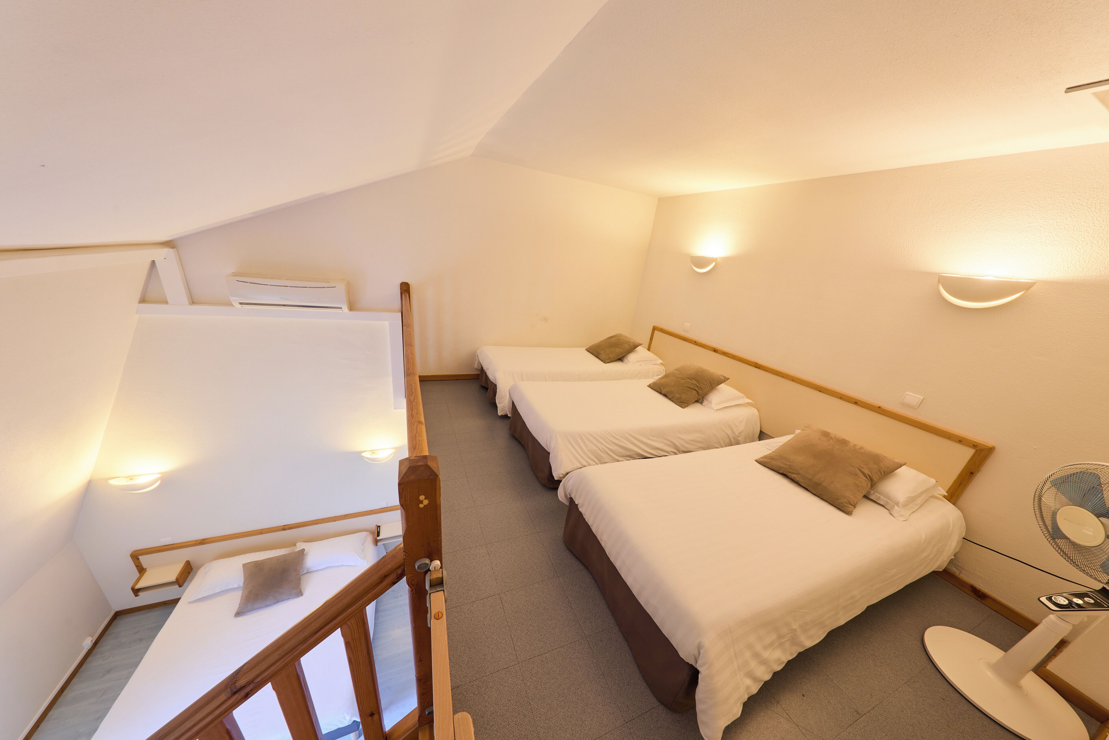 A dormitory-style room with three single beds, a small table with a fan, and a staircase leading to an upper level.