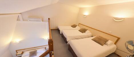 A dormitory-style room with three single beds, a small table with a fan, and a staircase leading to an upper level.