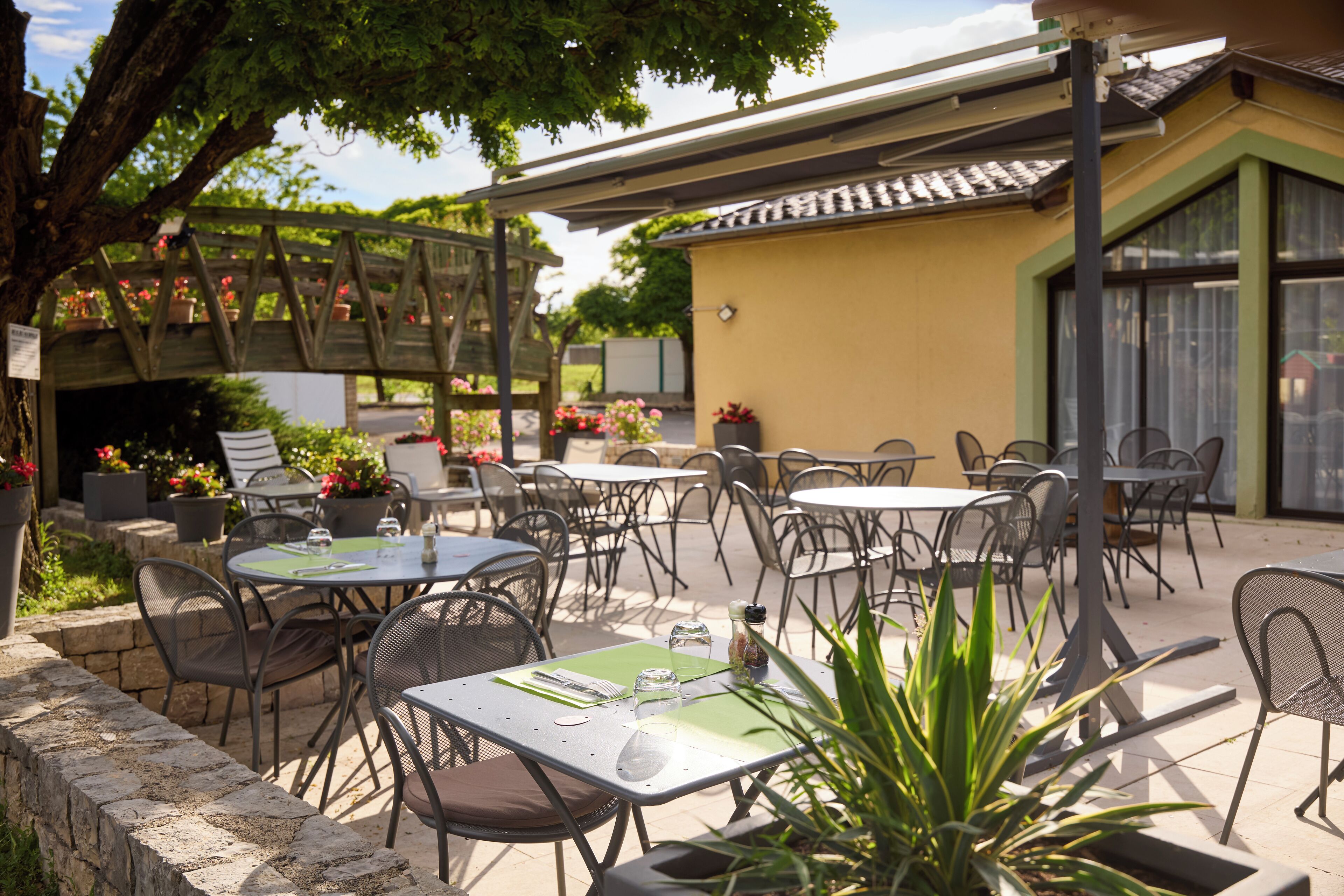 Un patio exterior con mesas y sillas, rodeado de plantas en macetas y un puente de madera.
