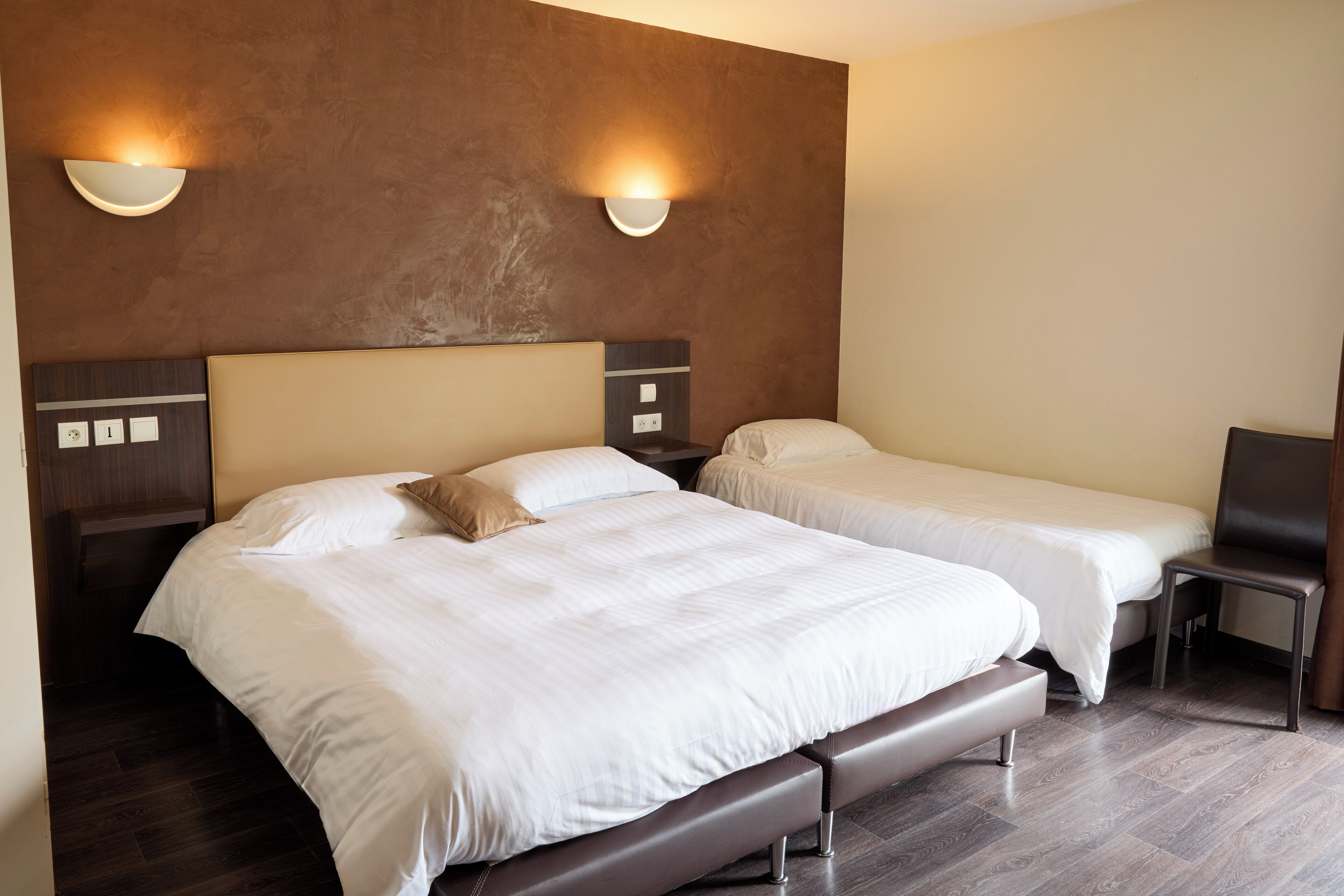 Habitación de hotel con dos camas, un banco, una silla y luces fijadas a la pared.
