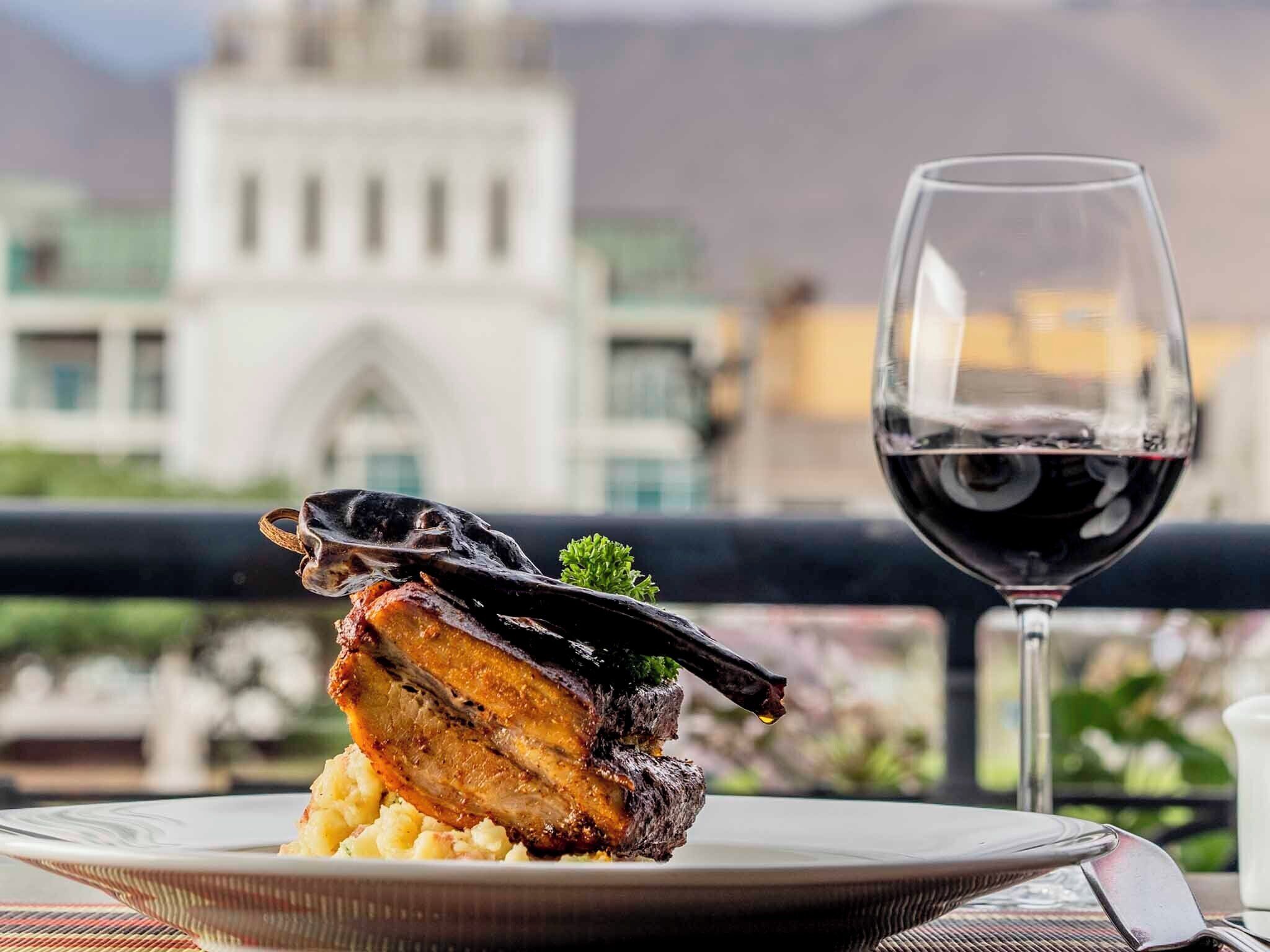 A plate of food with a glass of red wine on a balcony with a view of a building.