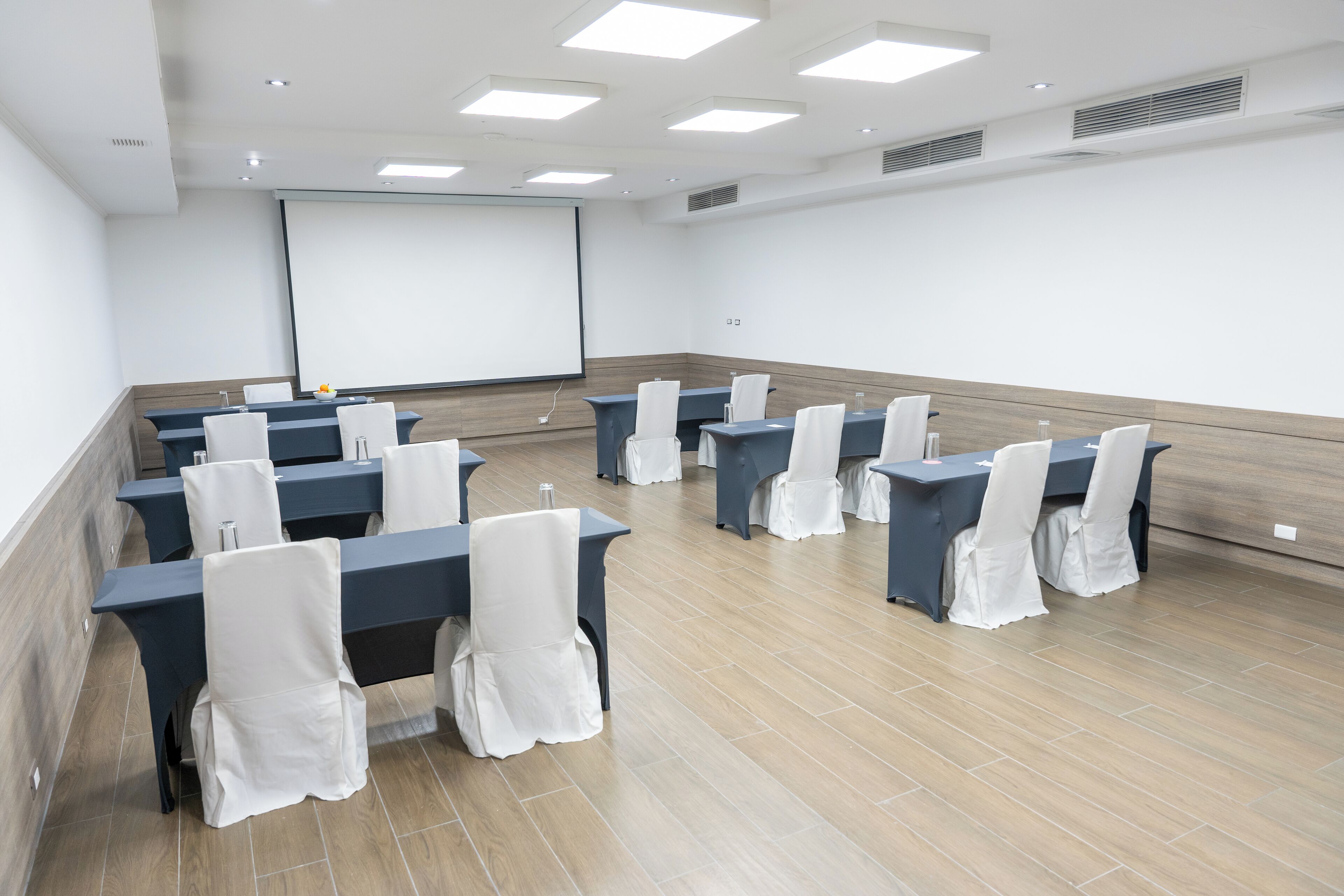Un salón de conferencias con varias mesas y sillas, una pantalla de proyector y una pizarra.