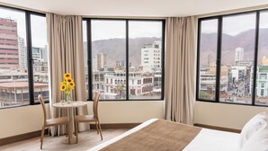 Quarto de hotel com uma janela grande, uma cama, uma mesa redonda com um vaso de girassóis e uma cadeira.