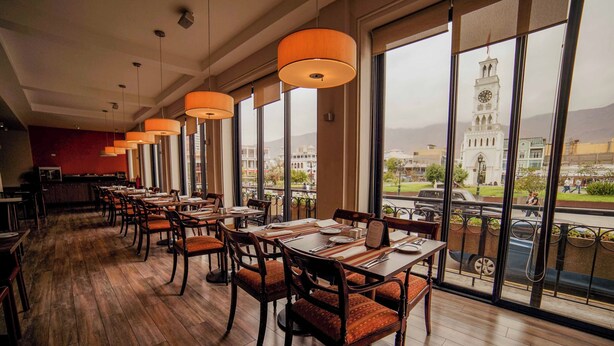Um restaurante com piso de madeira, cadeiras estofadas em vermelho e grandes janelas que oferecem vista para uma torre do relógio e paisagem urbana.