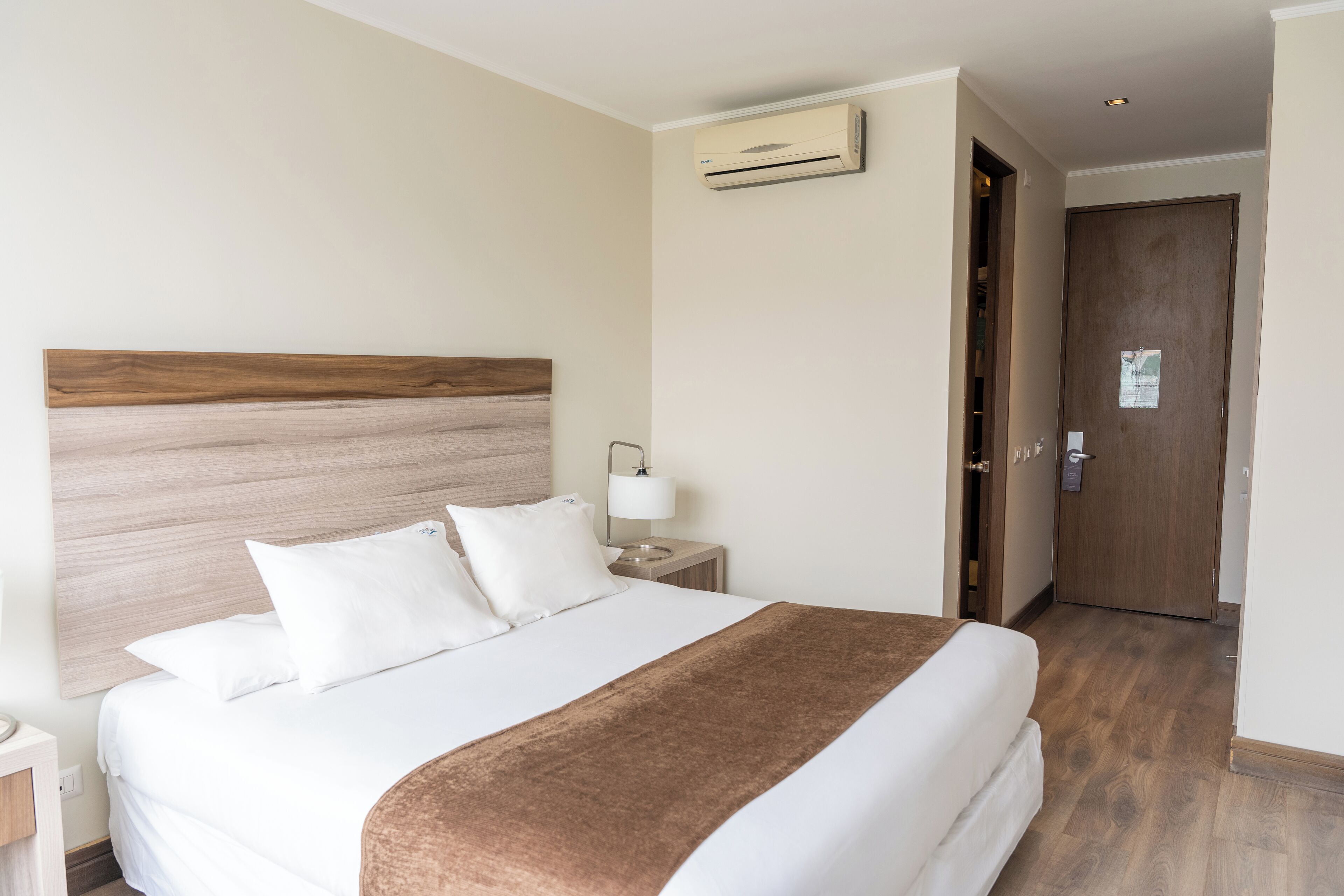 Una habitación de hotel con una cama grande, un cabecero de madera, una mesita de noche con una lámpara y una puerta que da a otra habitación.