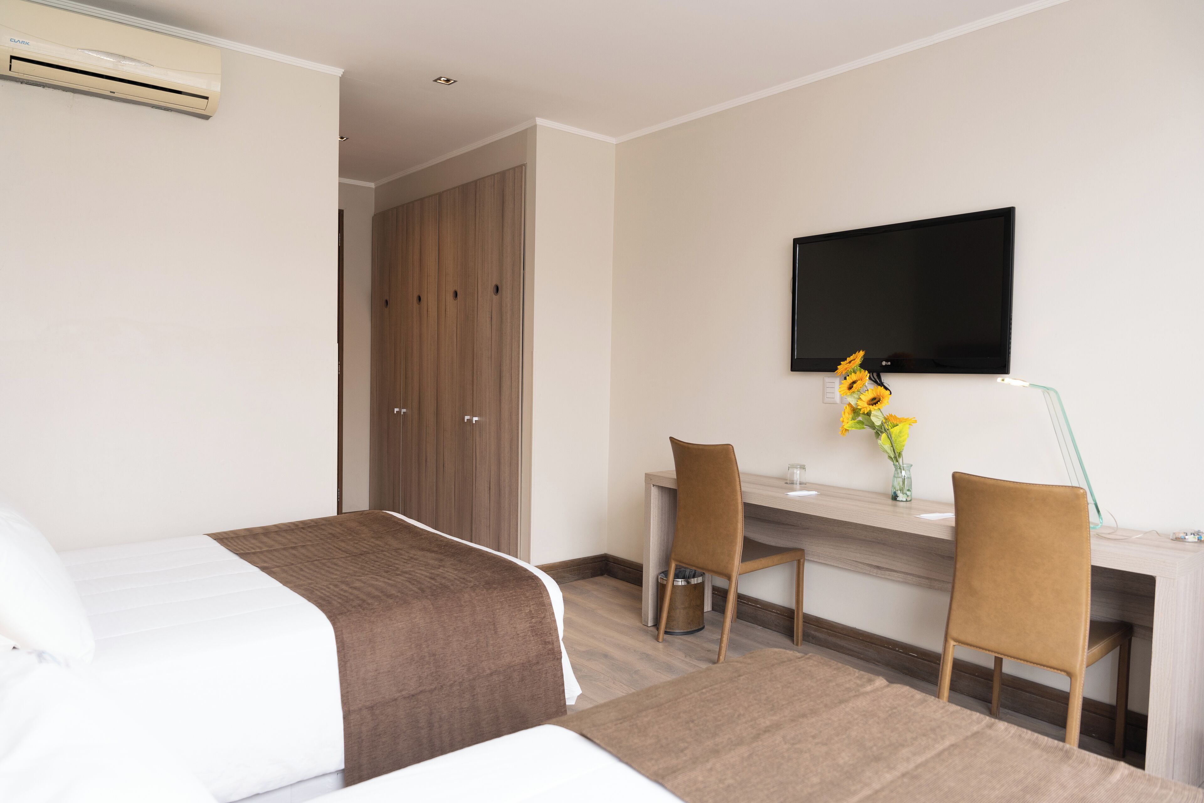 A hotel room with two beds, a desk with a chair, a flat-screen TV, and a vase with flowers.