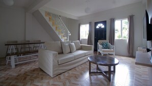 Un salon spacieux comprenant un canapé, une table basse, une table à manger avec des chaises, un escalier et une grande fenêtre avec des rideaux.