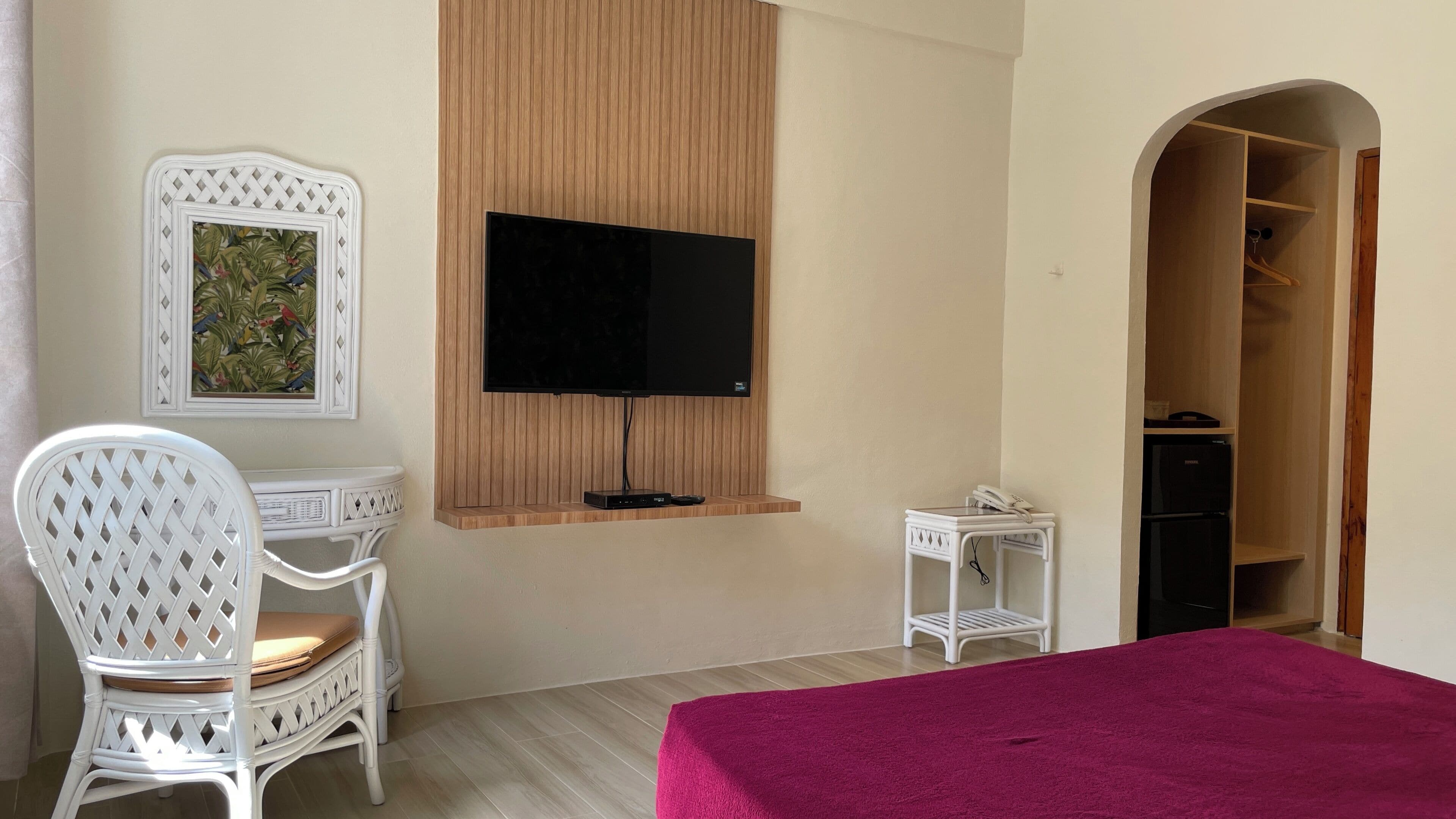 A hotel room with a bed, a desk with a chair, a television mounted on a wooden panel, a small table with a phone, and a wardrobe.