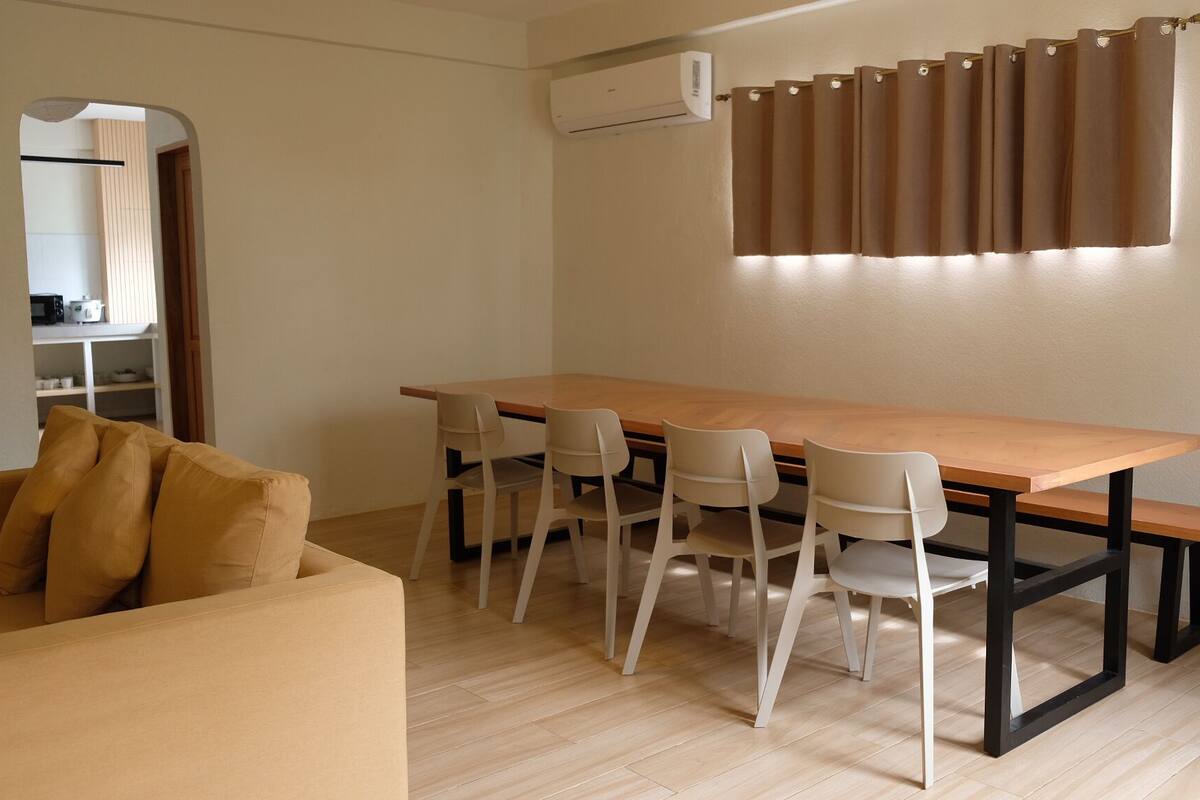 A compact living space with a wooden dining table, four chairs, a sofa with cushions, a kitchen area with a microwave, and an air conditioning unit.